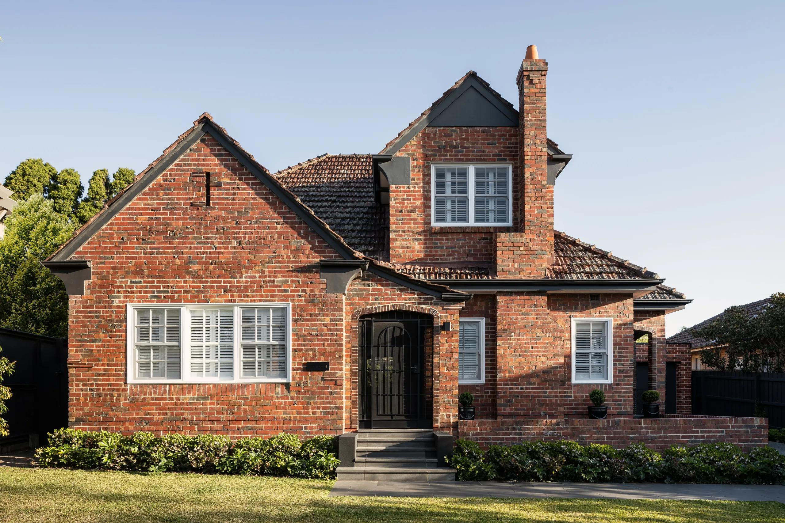 Red brick heritage frontage with tiled roof and chimney behind a clipped hedge